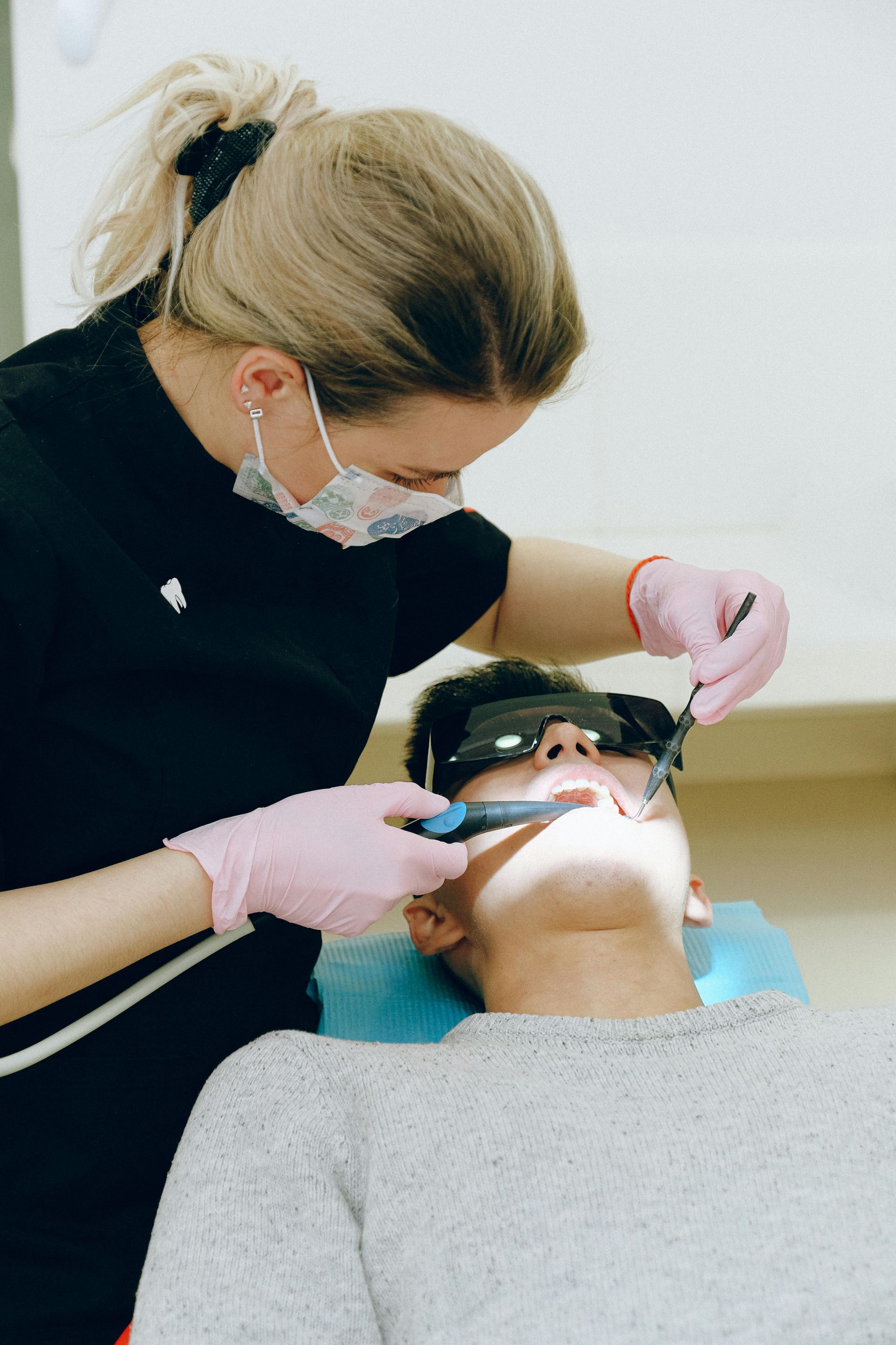 Dentist in mask and gloves examining a patient’s teeth with instruments. Patient wearing protective eyewear in dental chair.
