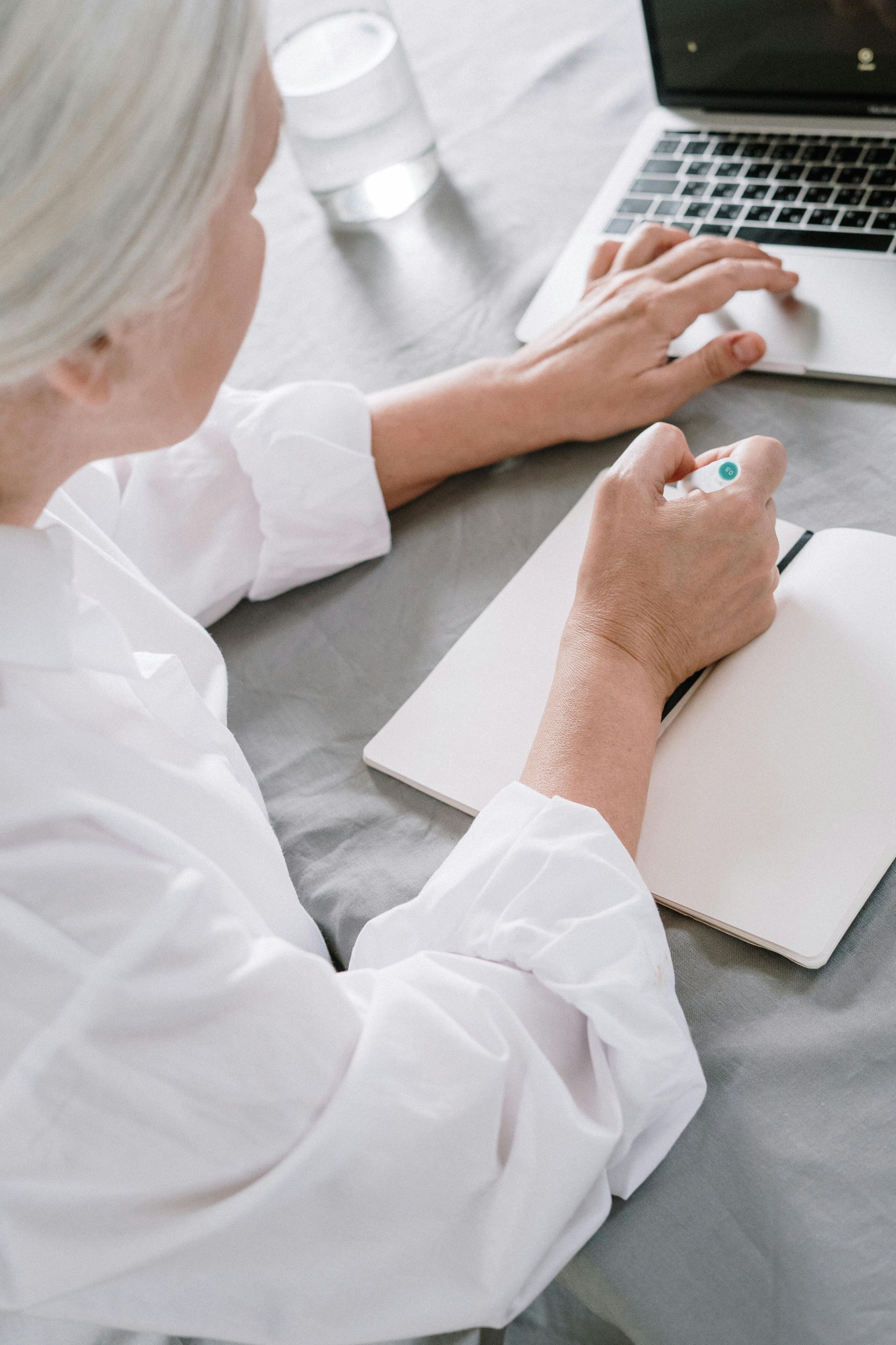 Person with white hair writing in a notebook, using a laptop, and holding a pen at a desk.