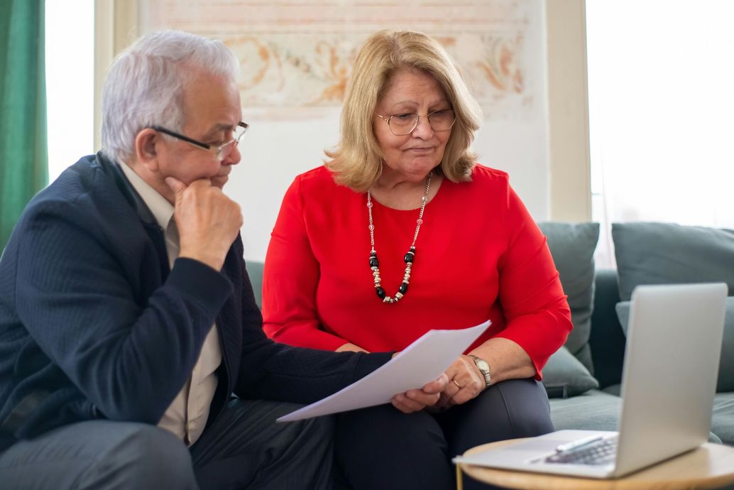 Older couple reviewing documents together, looking at laptop on a table in front of them.