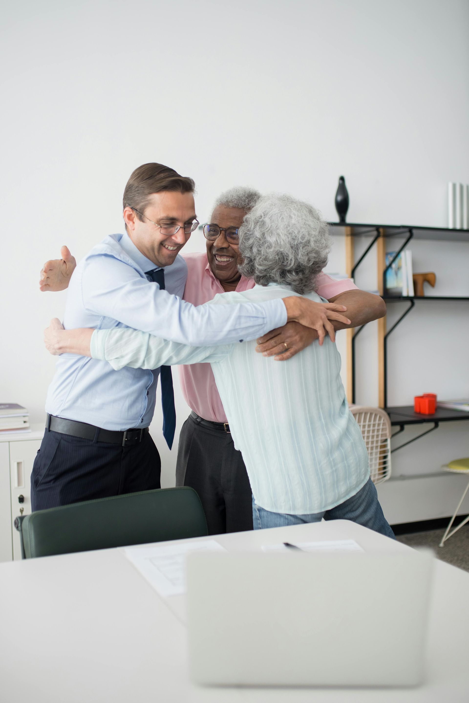 Three people in office embracing, smiling. Man in blue shirt, two older people. Laptop on desk.