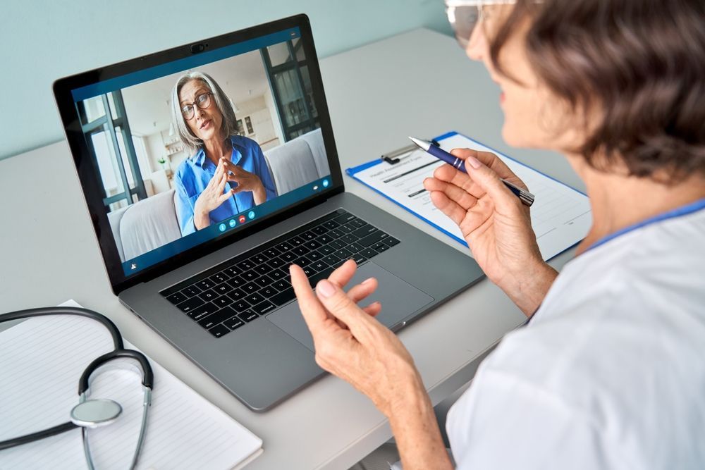 A healthcare provider in white scrubs on a video call with a patient; stethoscope and clipboard nearby.