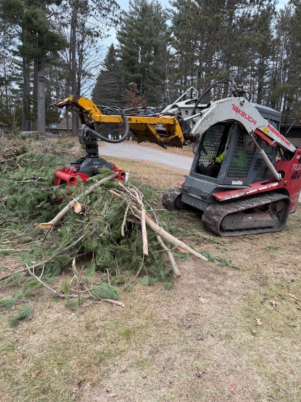 Skid steer and chipper processing tree debris in a wooded area. Red and yellow equipment, overcast day.