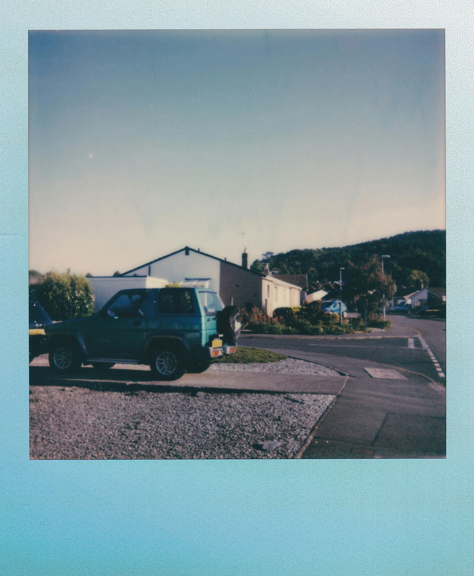 Green SUV parked on gravel driveway, houses and hills in background under clear sky.