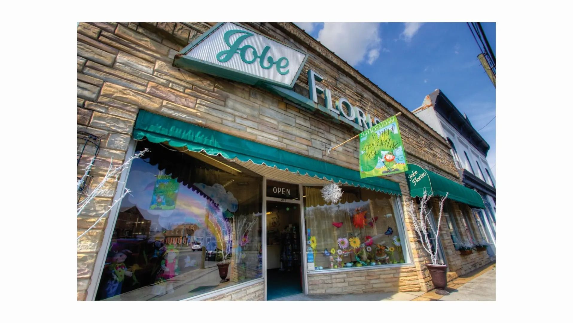 Jobe Flowers shopfront with teal awning, sign, and window display, on a brick building, under a blue sky.