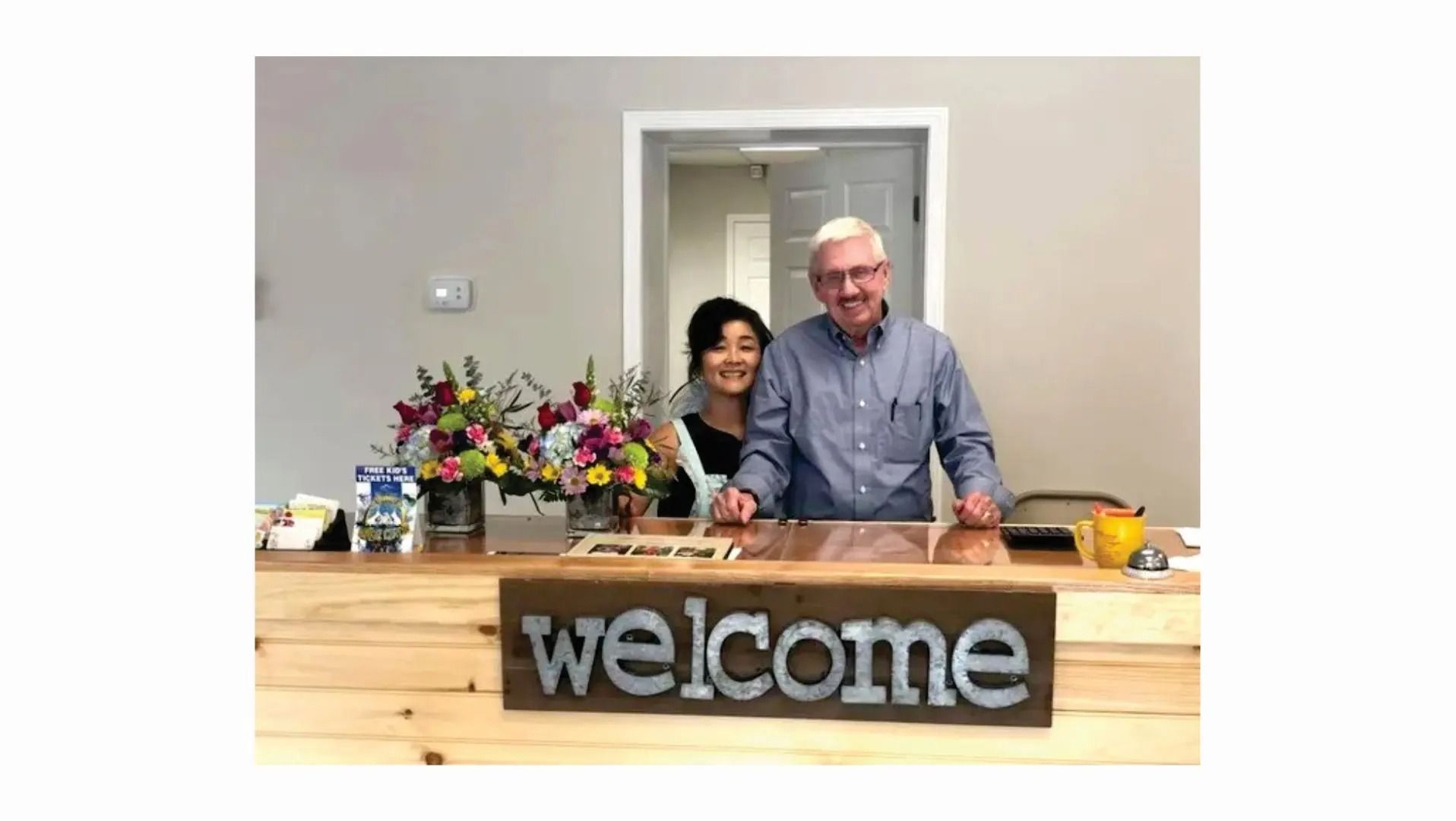 Two people smiling behind a wooden reception desk with a 