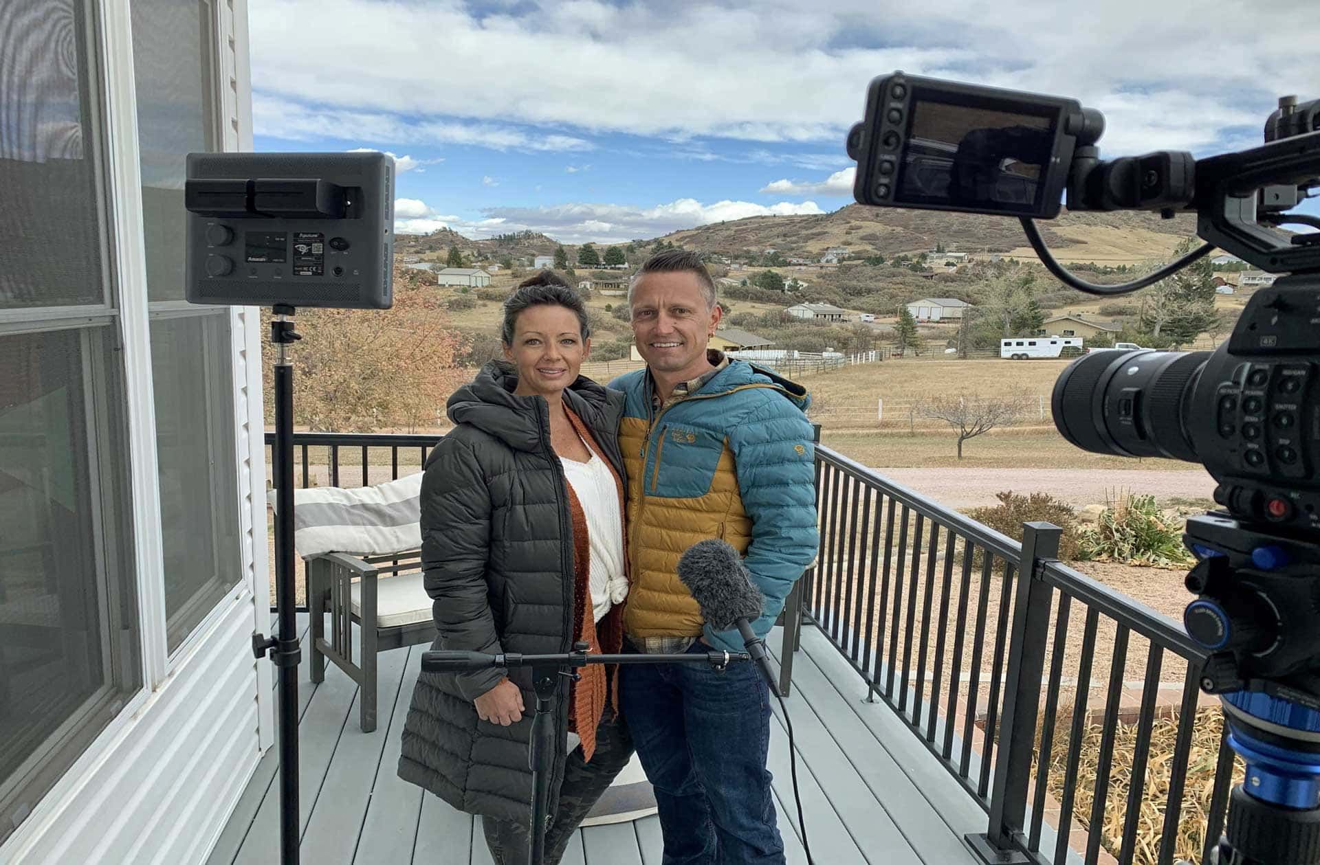 A man and a woman are standing on a deck in front of a camera.