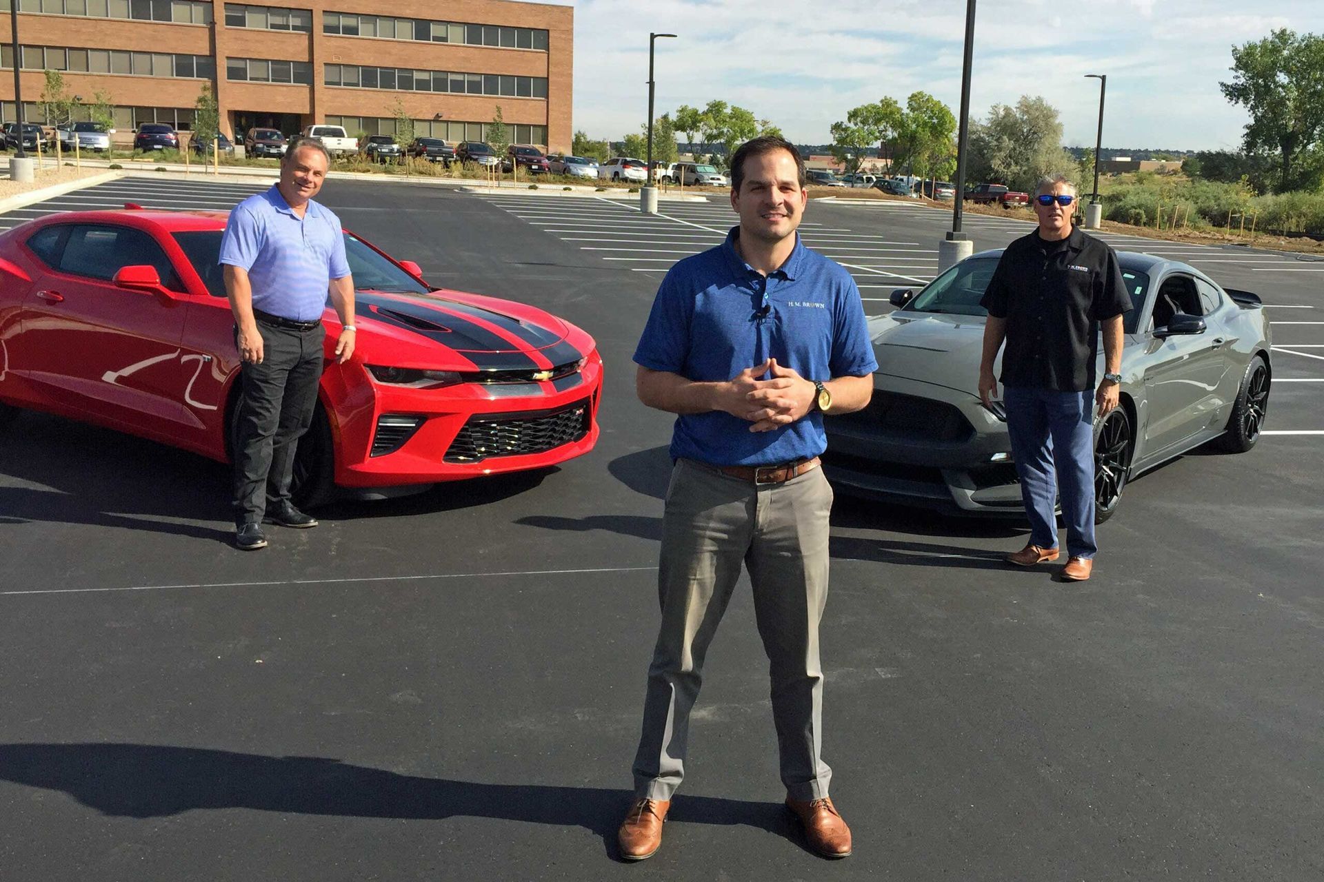 Three men are standing in front of two cars in a parking lot.