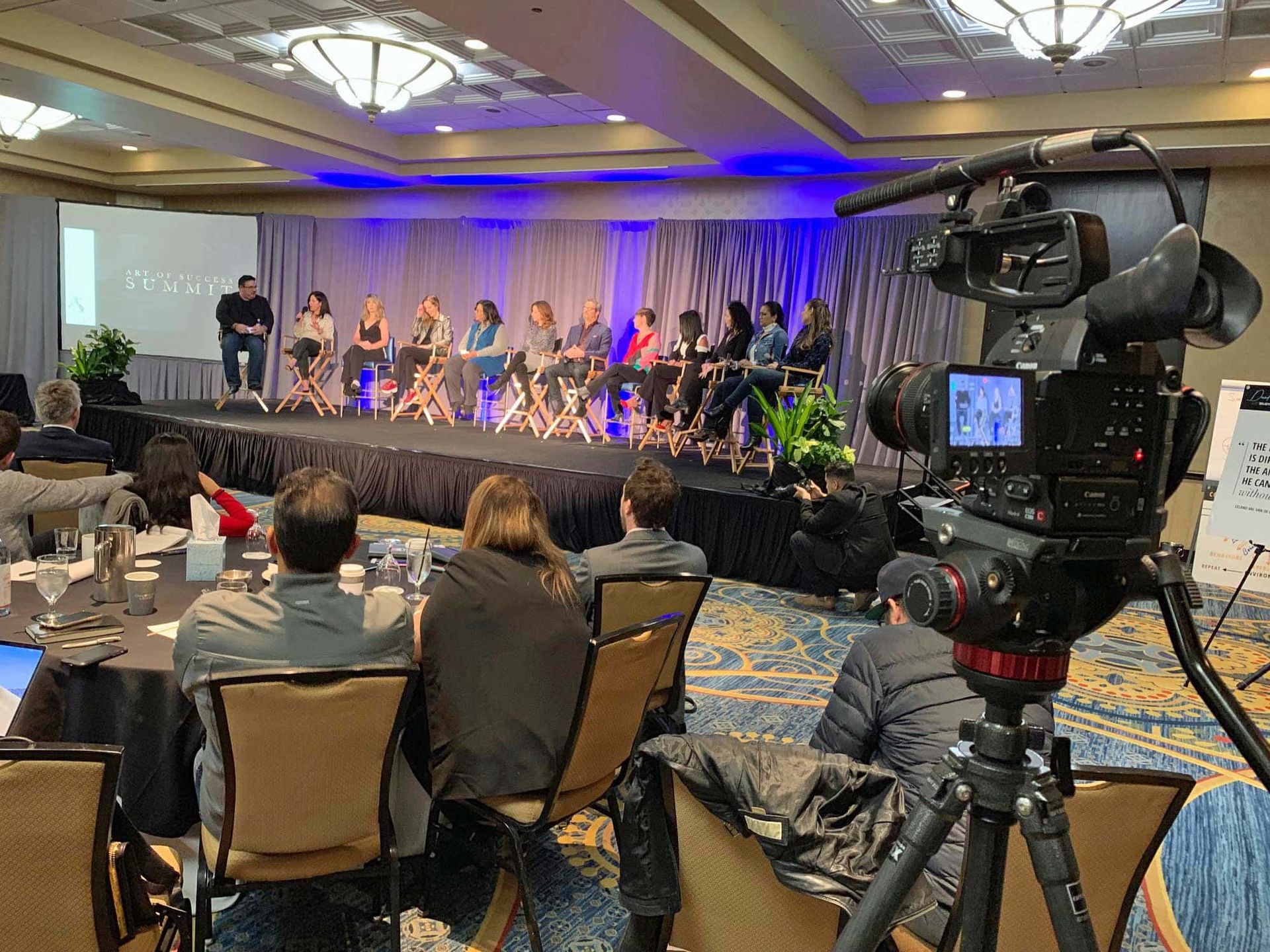 A group of people are sitting at tables in front of a stage with a camera.
