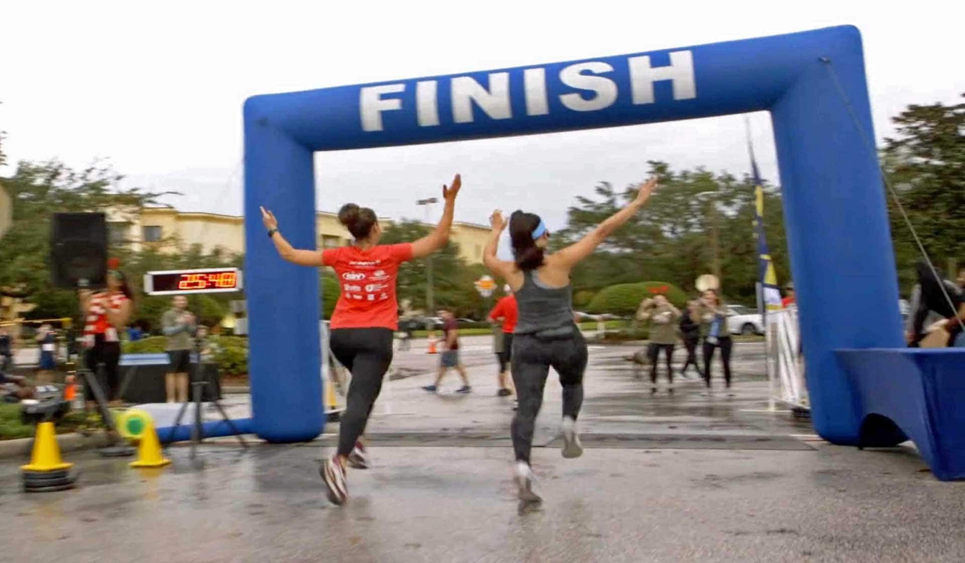 Two women are running under an inflatable finish line arch.