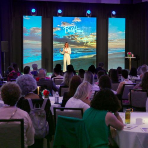 A group of people are sitting at tables in front of a screen that says baby house