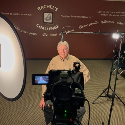 A man is kneeling in front of a camera with the words rachel 's challenge on the wall behind him