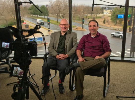 Two men are sitting in front of a camera in front of a window