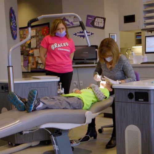 A woman in a pink shirt that says graces is standing next to a child in a dental chair