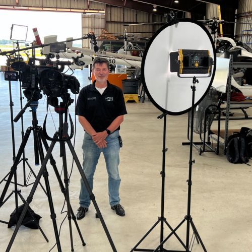 A man stands in front of cameras and lights in a hangar
