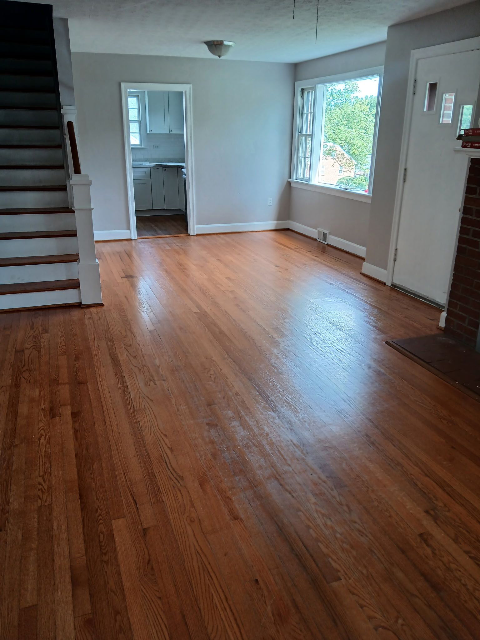 Hardwood floor in a living room with stairs, a doorway to a kitchen, and a window.