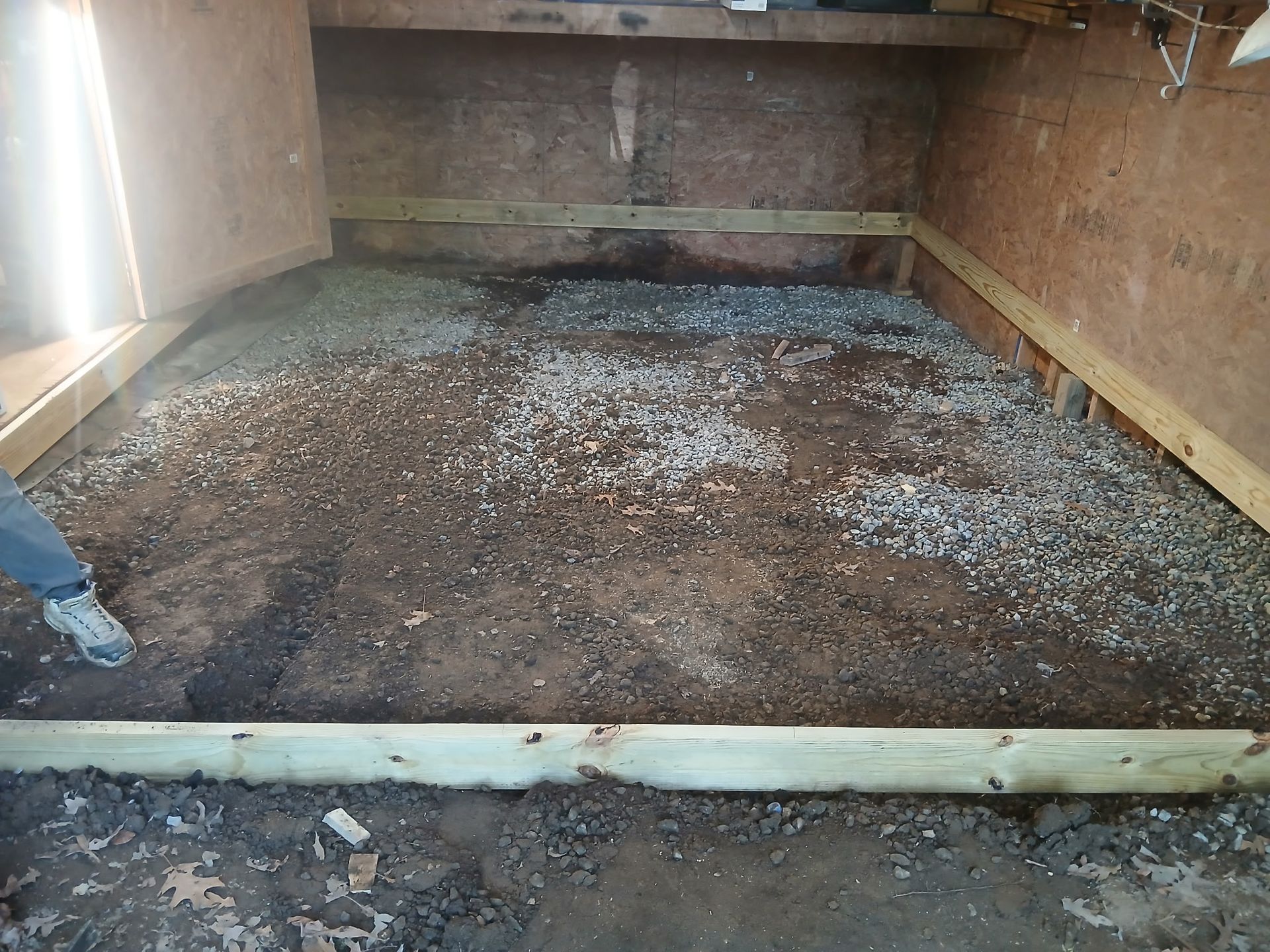 Gravel and soil fill a wooden-framed, ground-level area inside a dimly lit room. A person's leg and foot are in the left corner.