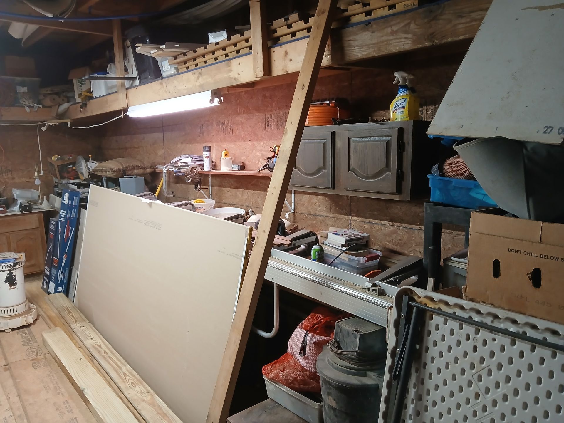 Cluttered garage with drywall, tools, and wood. Walls are wood and tan, with a fluorescent light and a fold-up table in the foreground.