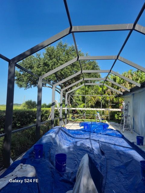 Screened patio under construction with metal frame, blue tarp covering floor, and trees visible beyond.
