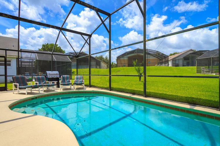 Screened backyard pool with turquoise water, patio chairs, and a green lawn under a blue sky