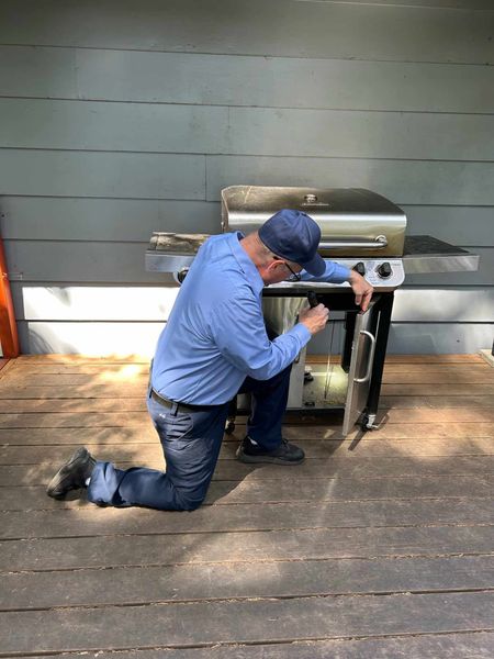 A man is kneeling down next to a grill on a deck.