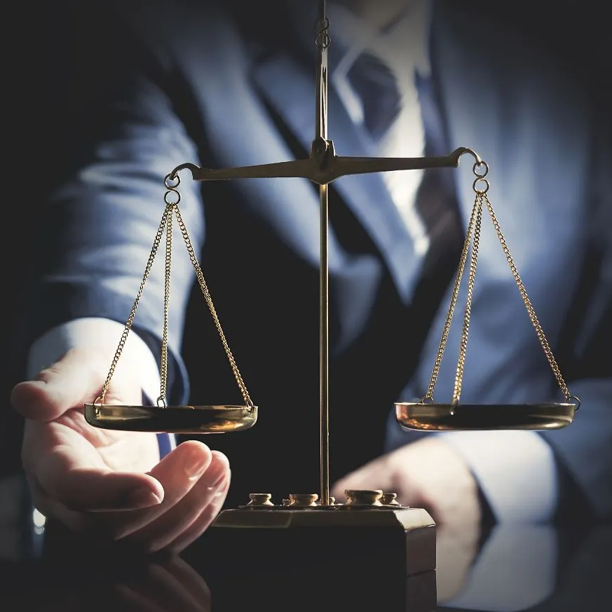 Hand holding balance scale, a symbol of justice, with a suited person in the background.