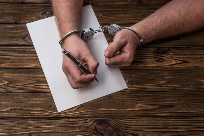 Hands in handcuffs writing on a blank paper with a pen, on a wooden surface.