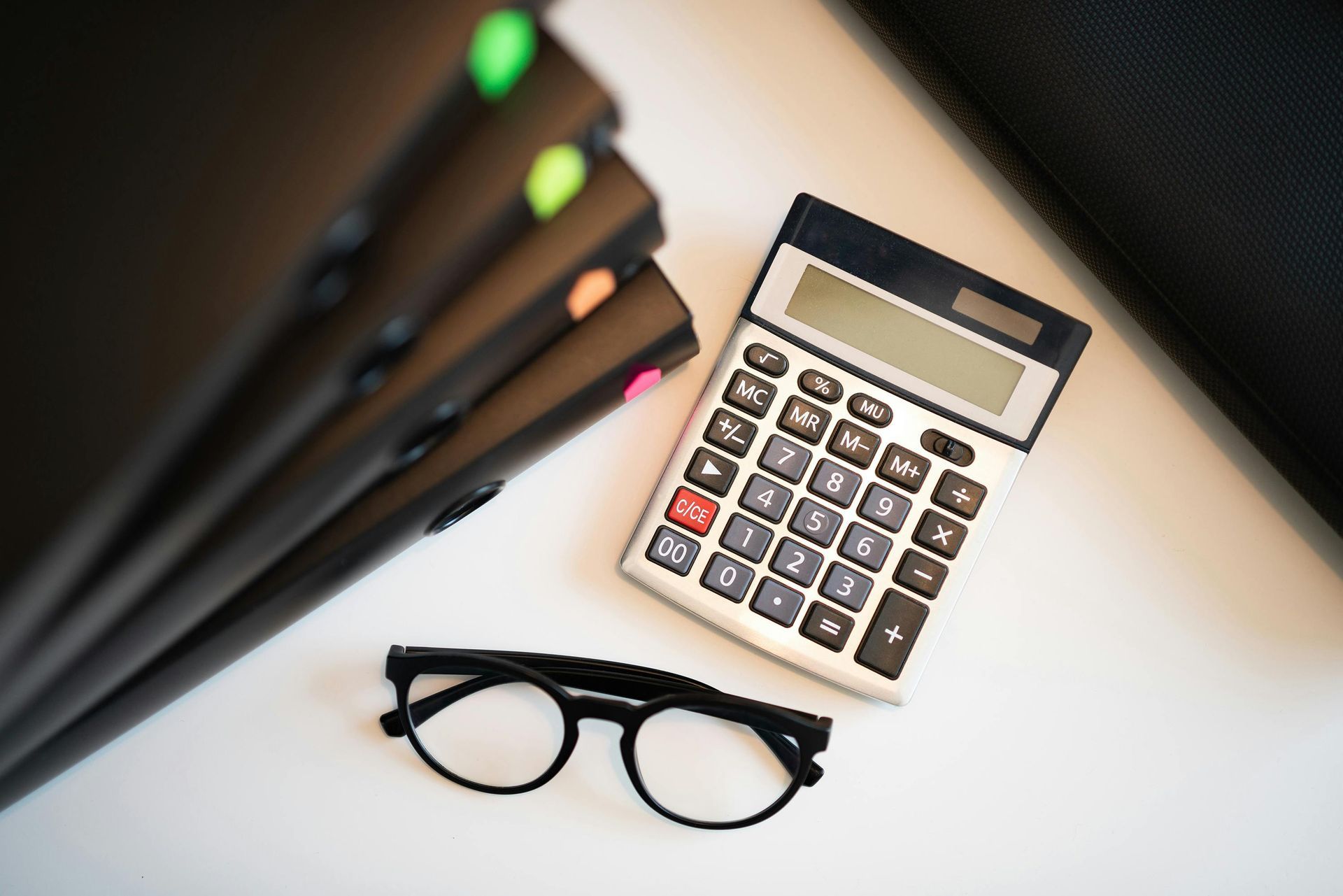 A calculator and glasses are on a table next to a stack of notebooks.