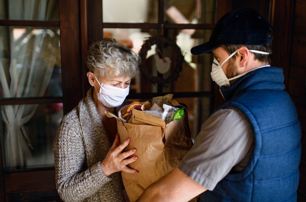A man wearing a mask is giving a bag of food to an elderly woman.