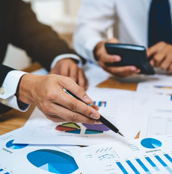 A man is pointing at a graph on a piece of paper while another man holds a cell phone.