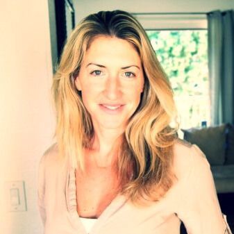 Woman with blonde hair smiles indoors, lit by natural light.