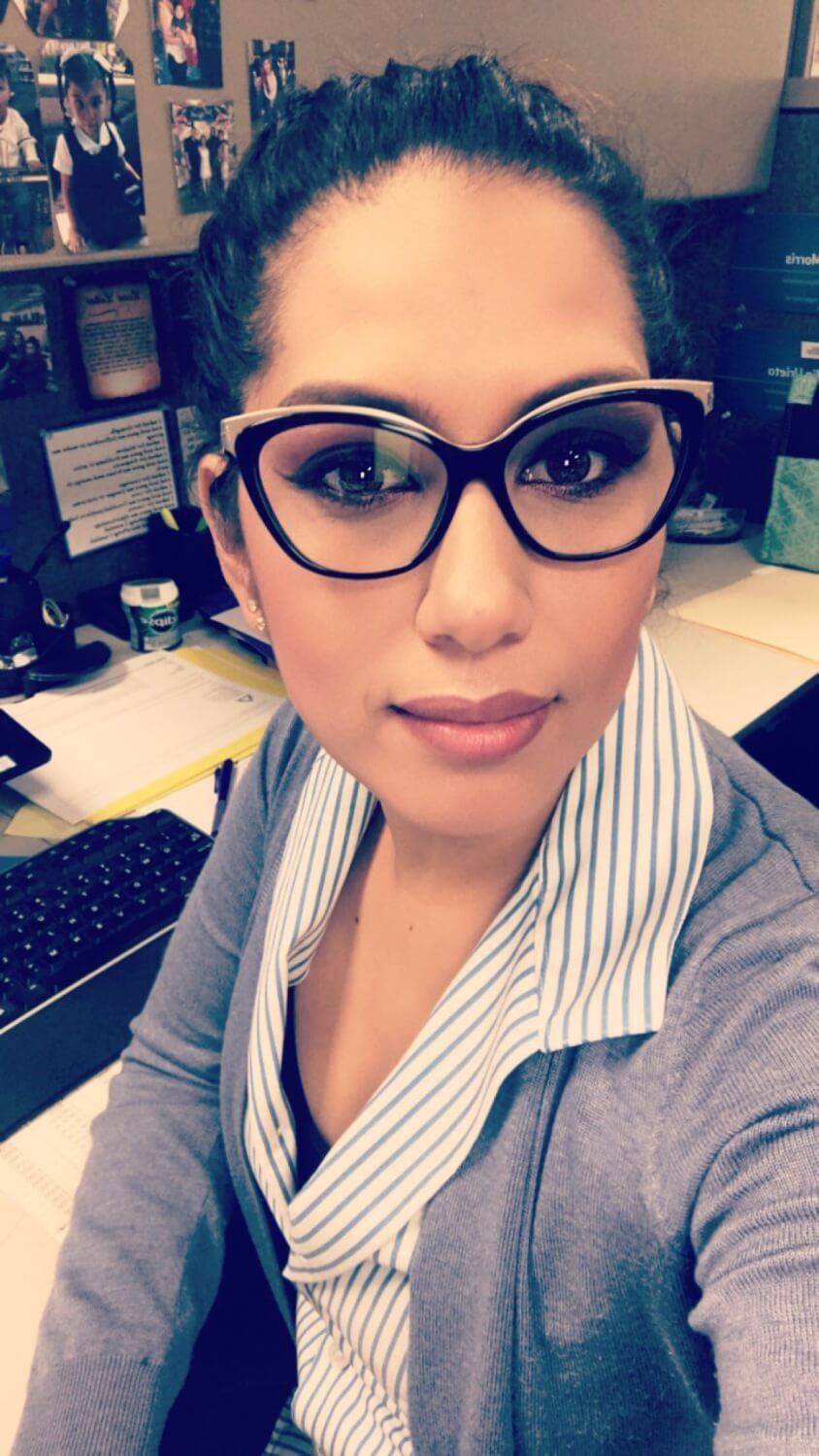 Woman wearing glasses, striped shirt, and gray cardigan, seated at a desk.