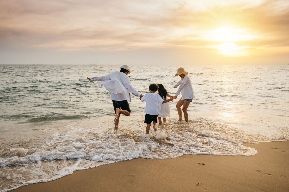 Family playing in the ocean at sunset; adults and children hold hands, wading in the water.