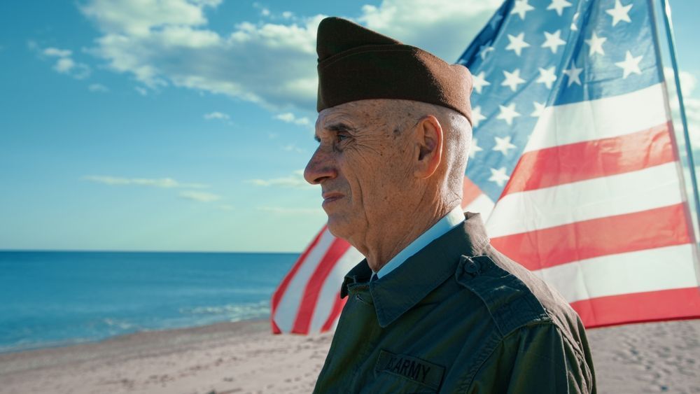 Veteran in uniform stands on beach with American flag in background, looking toward sea.