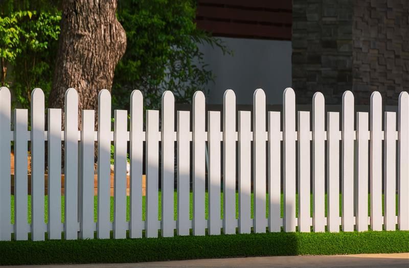 White picket fence in front of green grass, tree, and building.