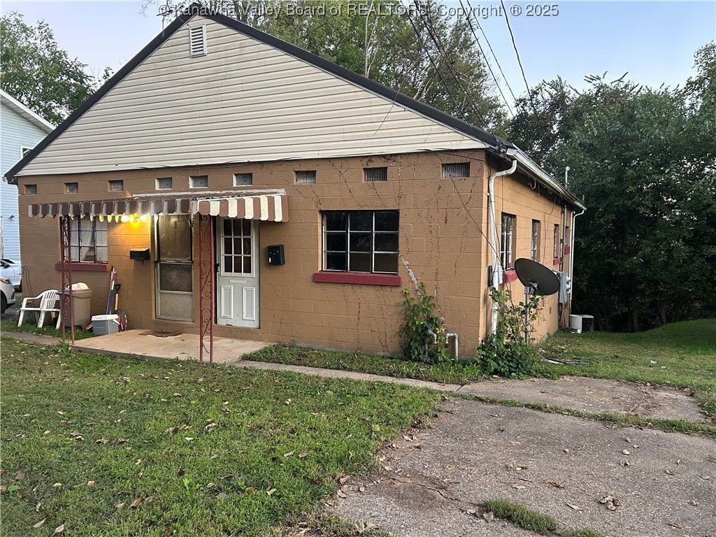 Tan stucco house with awning and small yard, driveway and trees in the background.