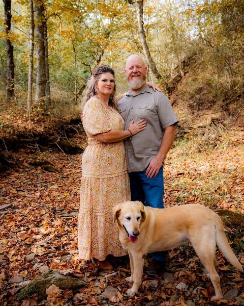 Couple with dog in a forest setting; woman in floral dress, man in shirt and jeans, dog is yellow lab, autumn leaves.