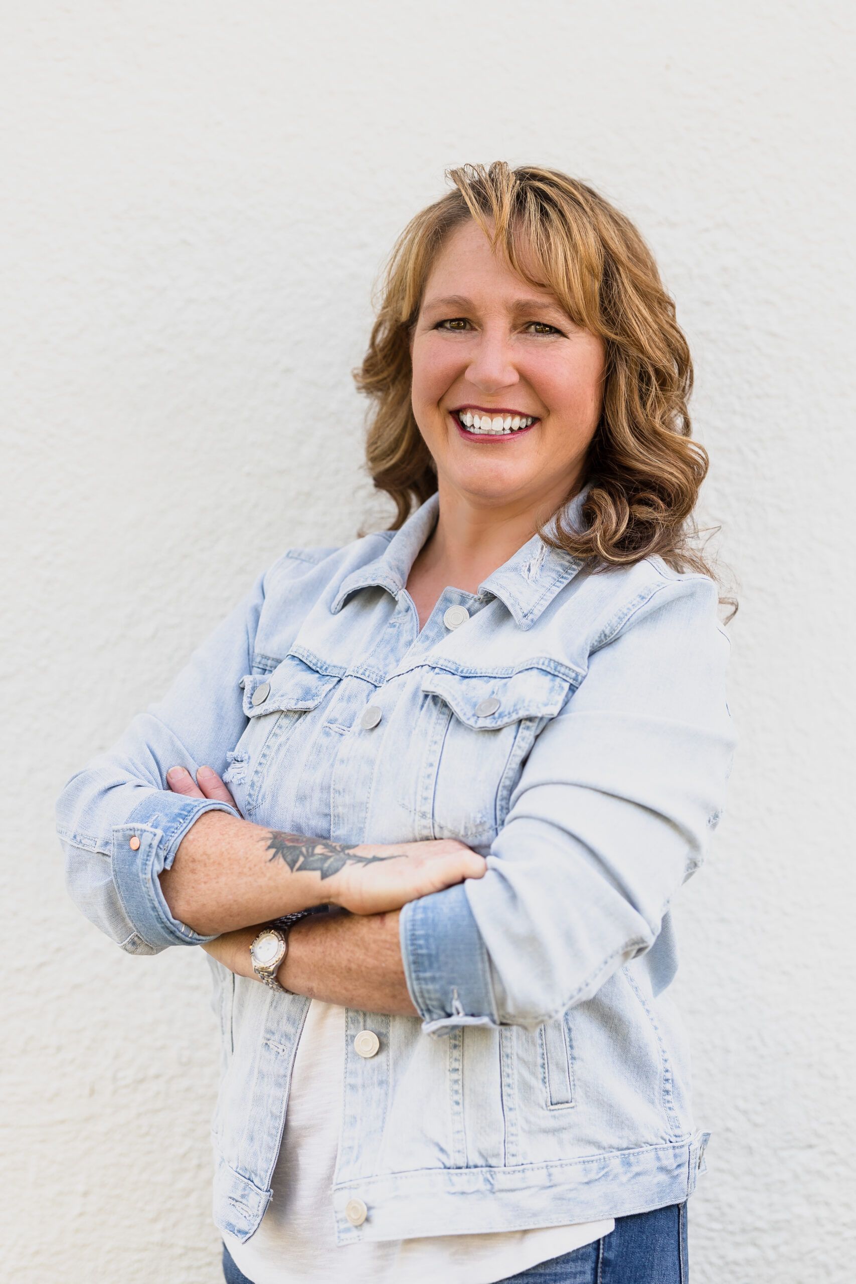 Woman with crossed arms smiles against a white wall, wearing a light-wash denim jacket.