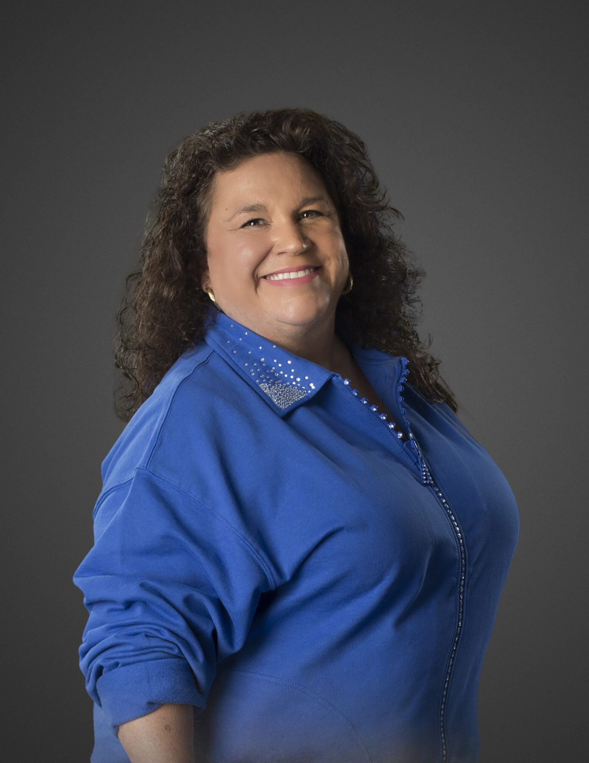 Woman in blue shirt smiling, posed against a gray backdrop.
