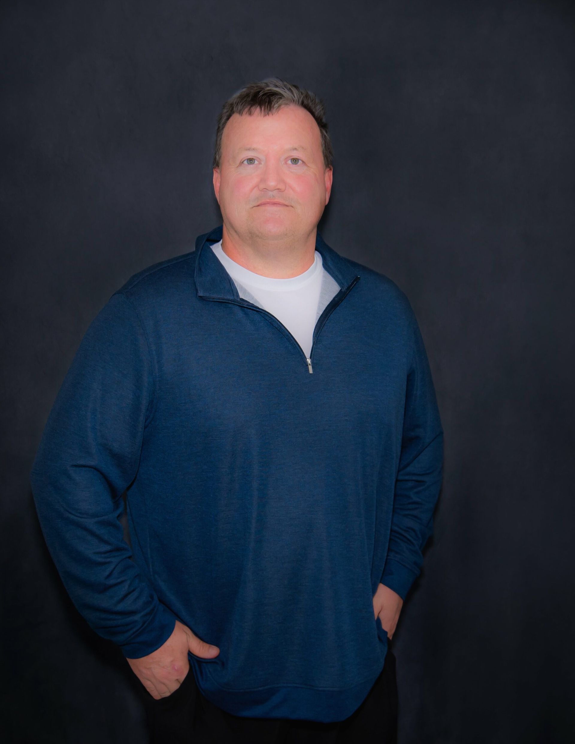 Man in blue sweater, hands in pockets, looking at the camera against a dark gray background.