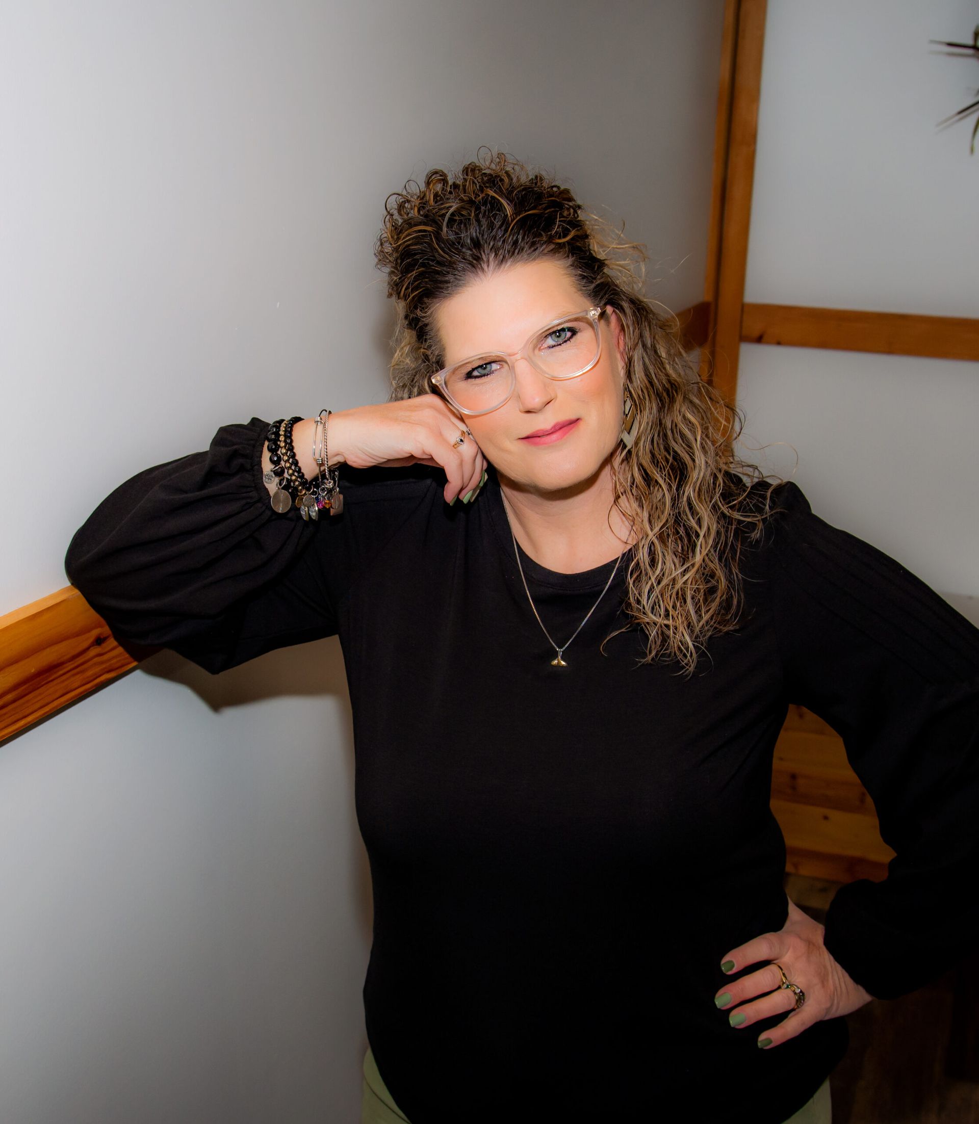 Woman with curly hair and glasses leans against a wall, resting arm on railing, and hand on hip.