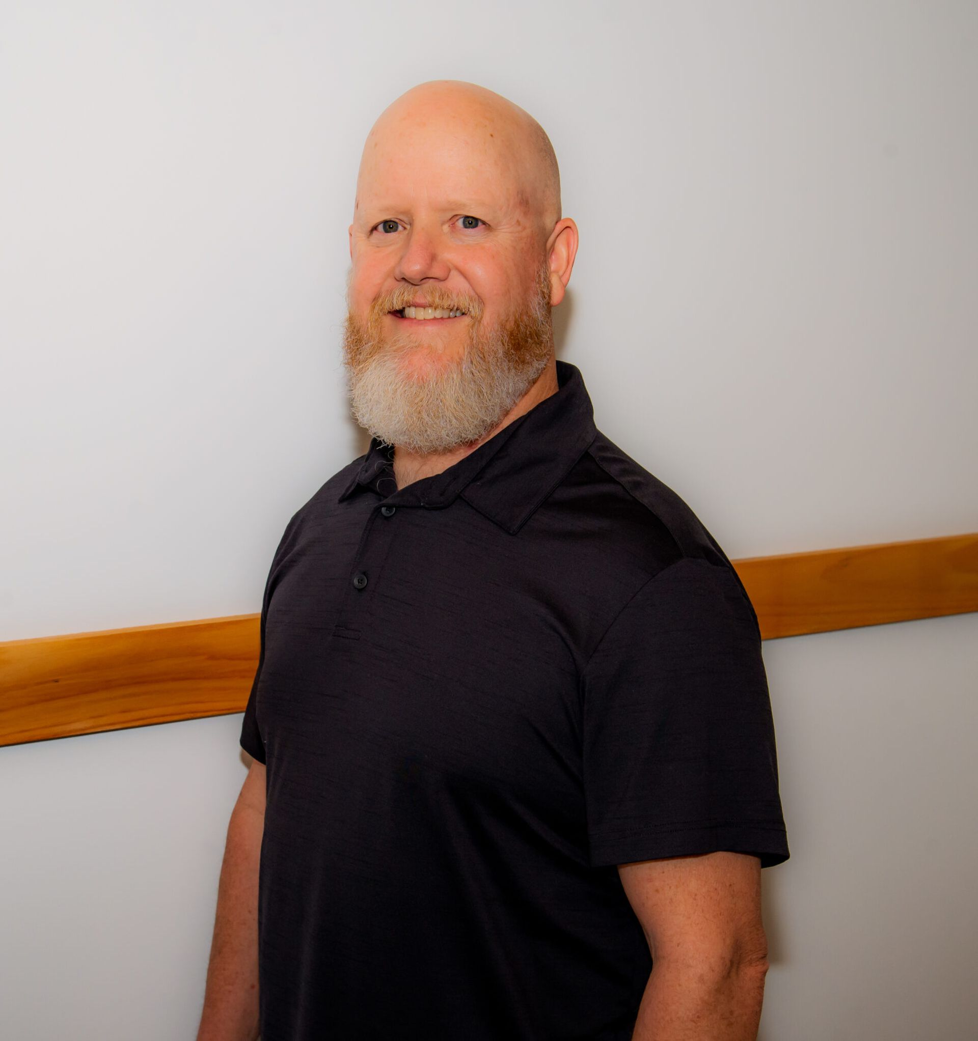 Bald man with a gray beard smiles, wearing a dark blue polo shirt, standing by a wall.