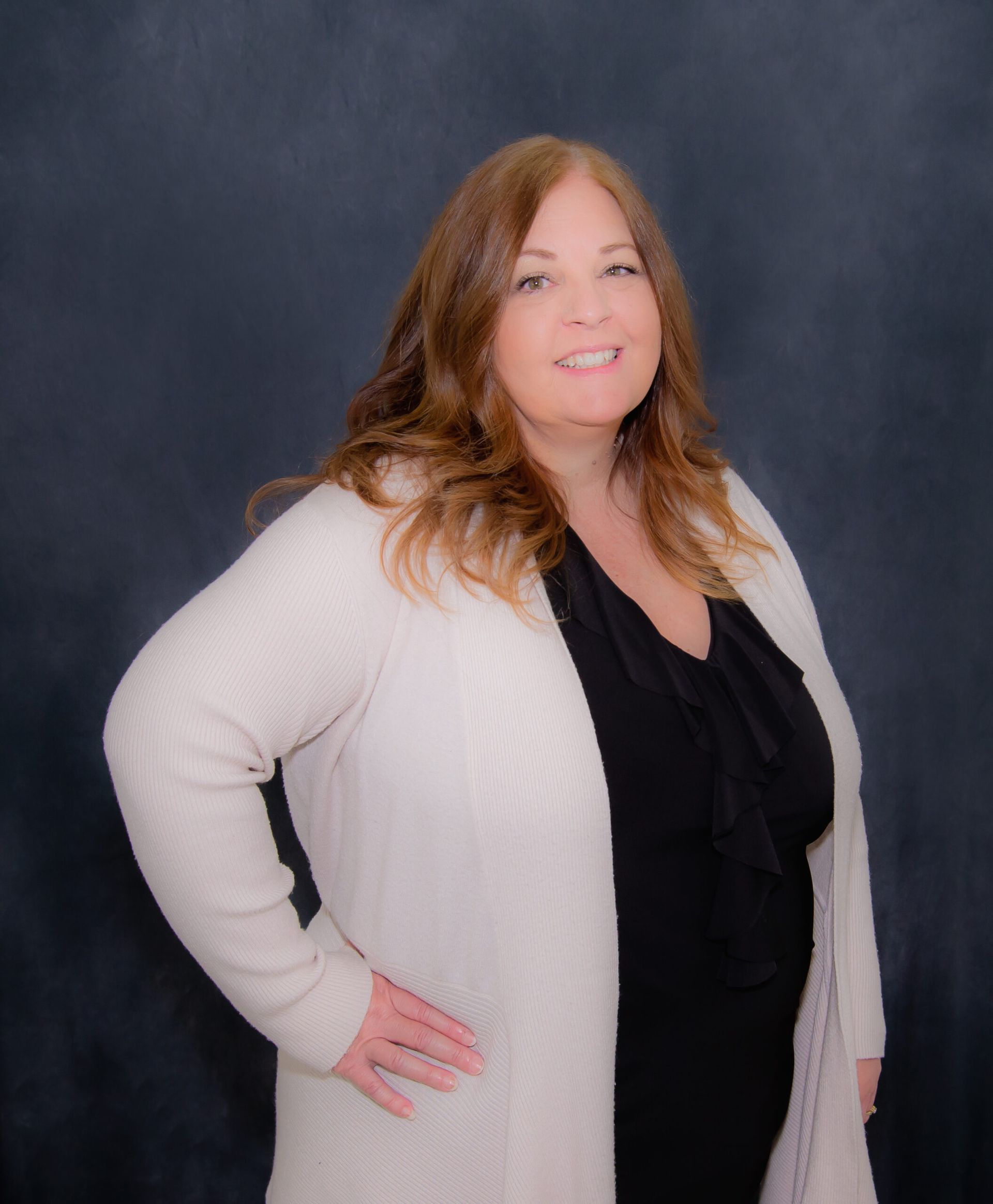 Woman with brown hair smiles, wearing a cream-colored sweater over a black top, poses against a dark backdrop.