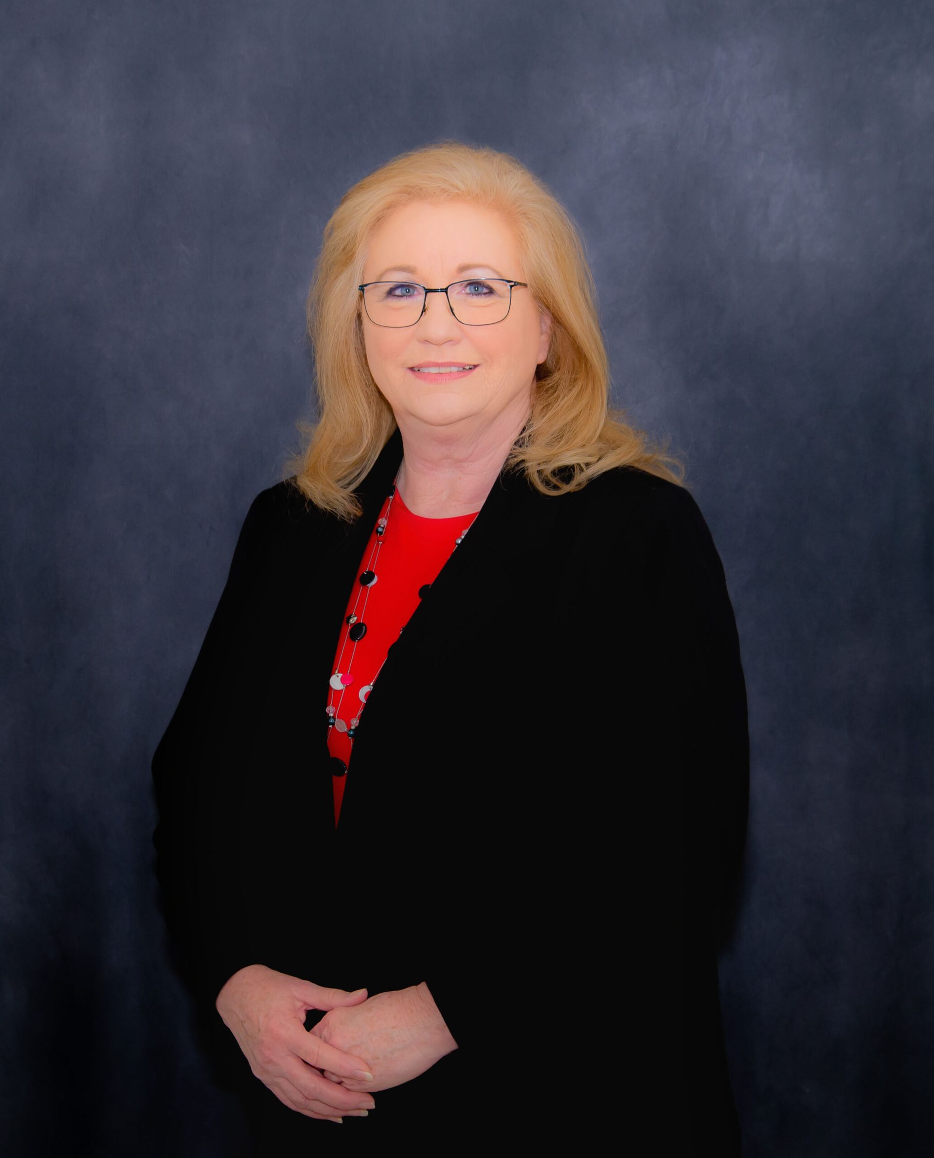 Woman with blonde hair wearing glasses and a black jacket, red top, arms crossed, on a blue background.