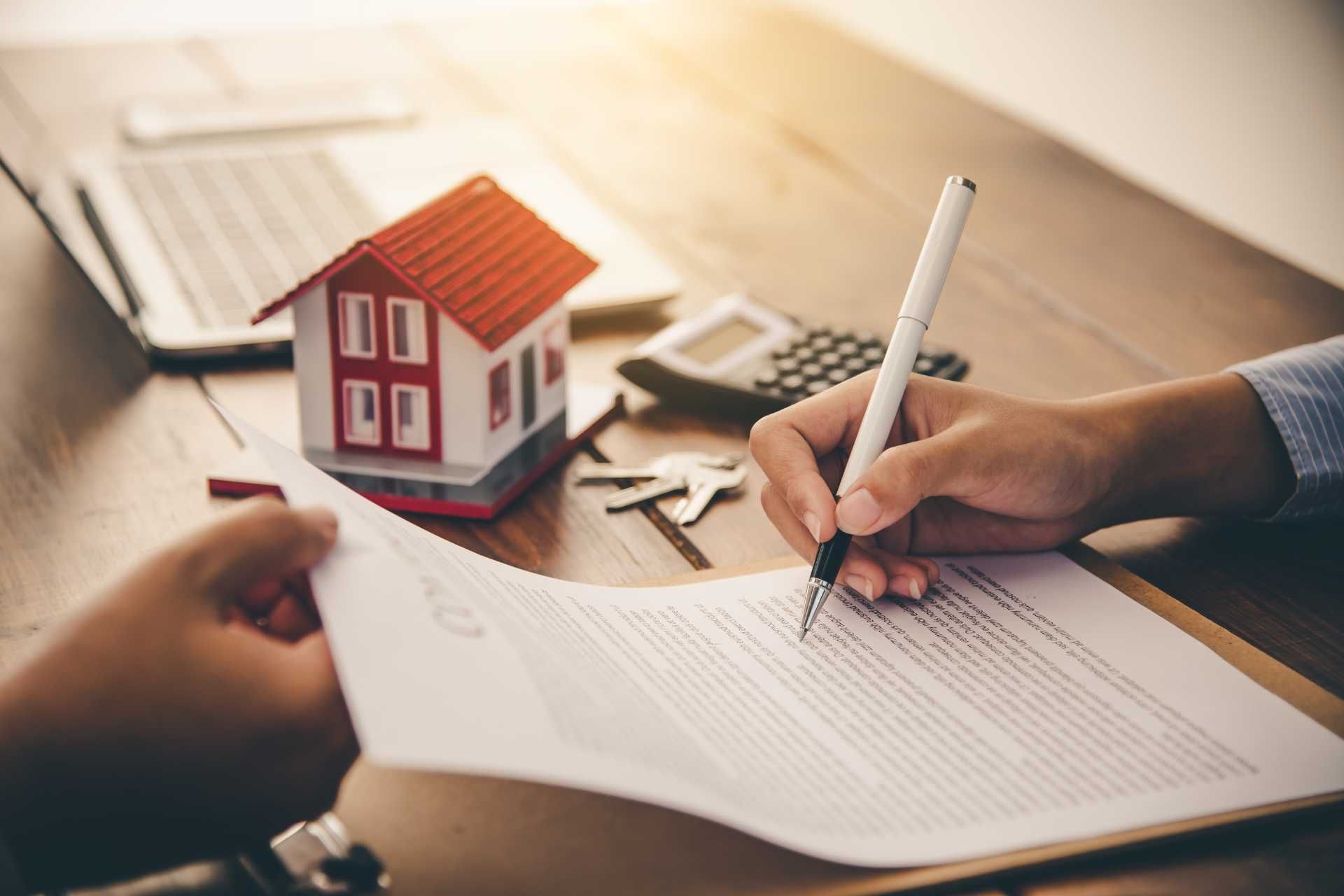 Person signing a real estate contract with a model house, keys, and calculator on a desk.