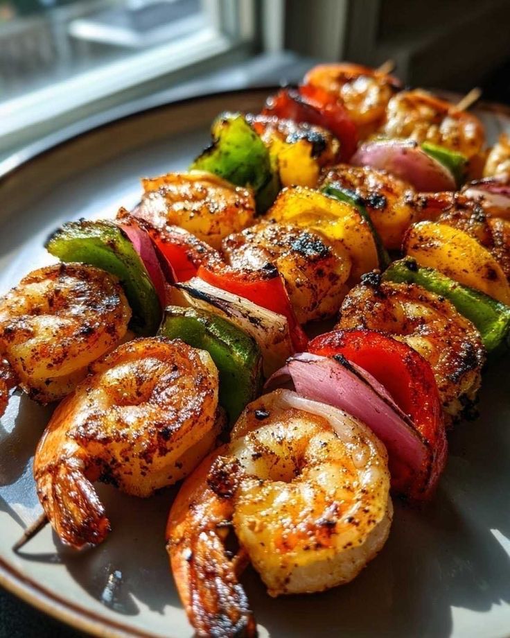 Grilled shrimp skewers featuring red onion, green, yellow, and red bell peppers on a plate near a sunny window.