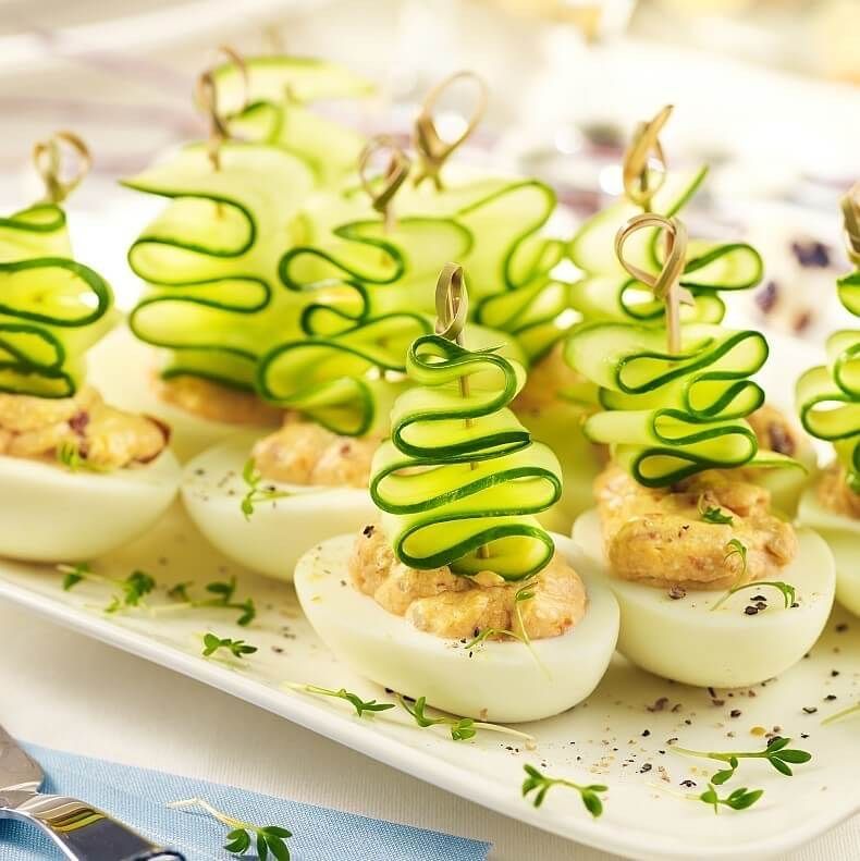 Deviled eggs topped with accordion-folded cucumber ribbons on cocktail picks, arranged on a serving tray with herbs.
