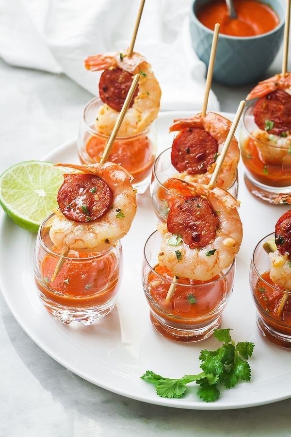 Skewered shrimp and chorizo served in small glasses with red sauce, garnished with a lime wedge and cilantro on a plate.