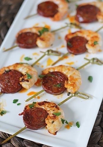 Skewers of cooked shrimp and seared chorizo slices on a white platter, garnished with fresh green herbs.