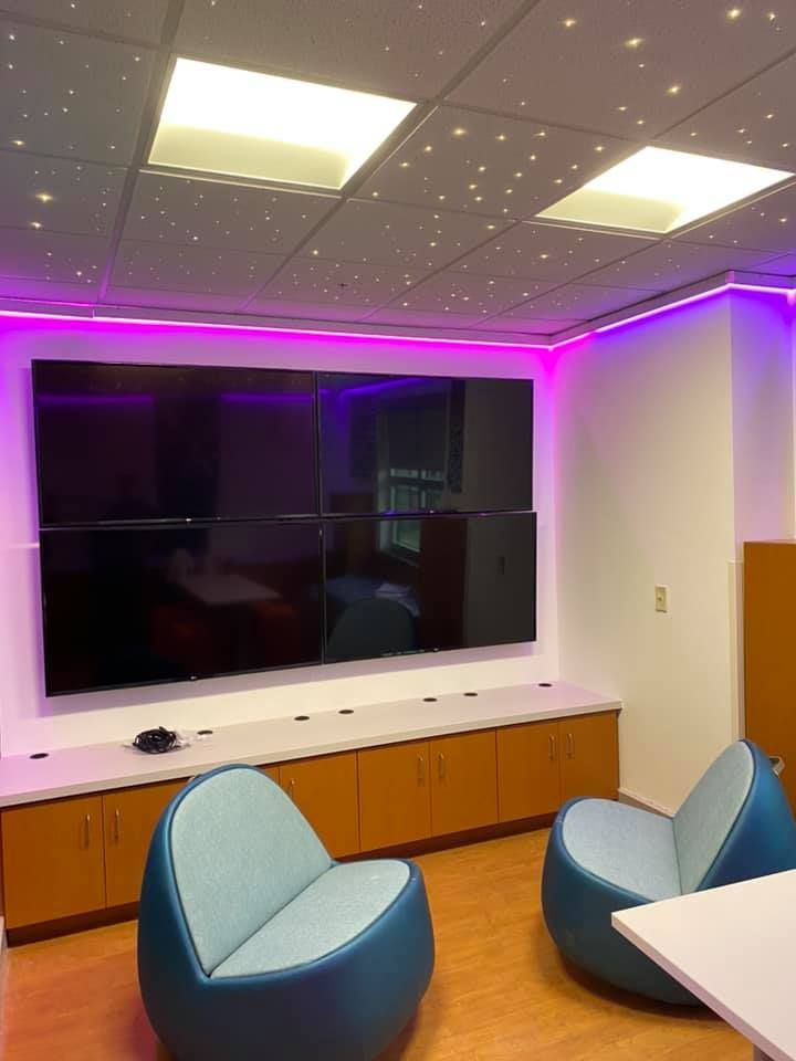 Room with six large black screens, purple LED lighting, and two blue chairs.