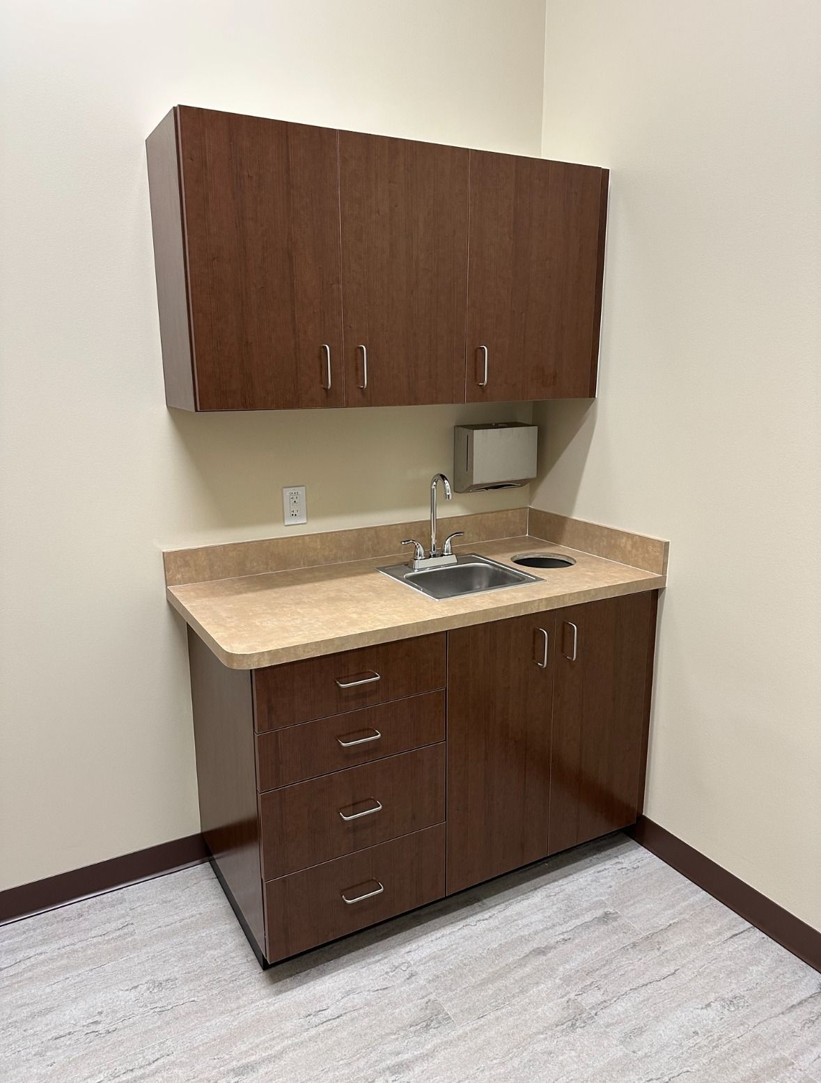 Medical cabinet with sink and overhead storage, brown cabinets, beige countertop, and light flooring.